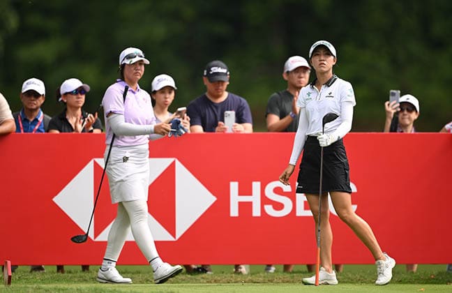 Two women are playing golf, with one holding a club and the other holding a cell phone. They are surrounded by a crowd of people watching the game. AI generated content
